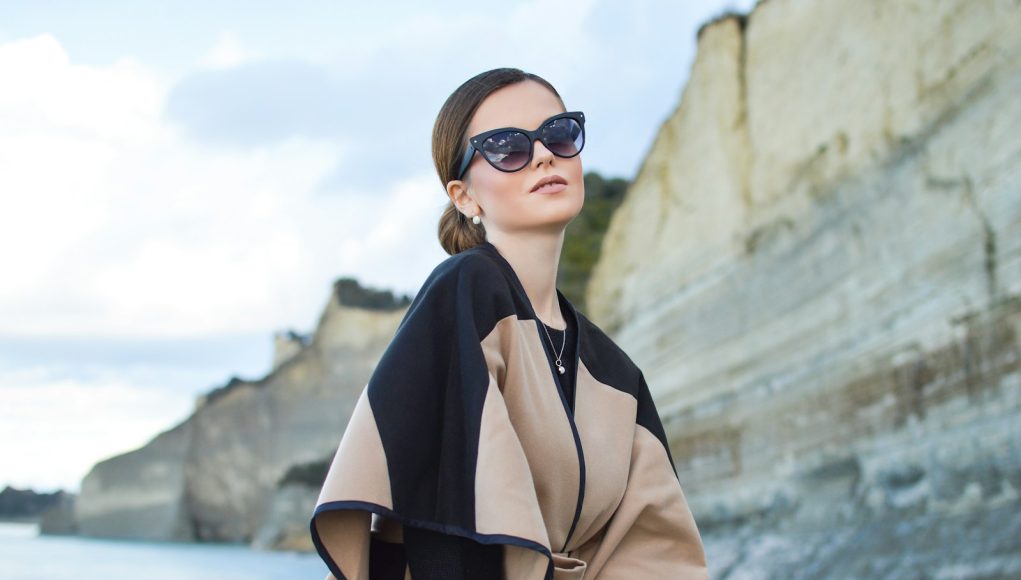woman wearing black and brown dress and sunglasses standing near body of water under white and blue sky